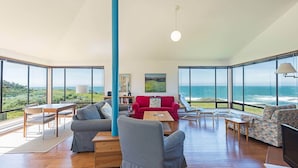 A spacious living room with a large wooden table, two sofas, and a red armchair. The room features large windows offering ocean views.