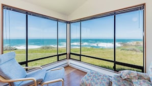 A room with large windows overlooking the ocean, featuring a blue chair and a patterned ottoman.