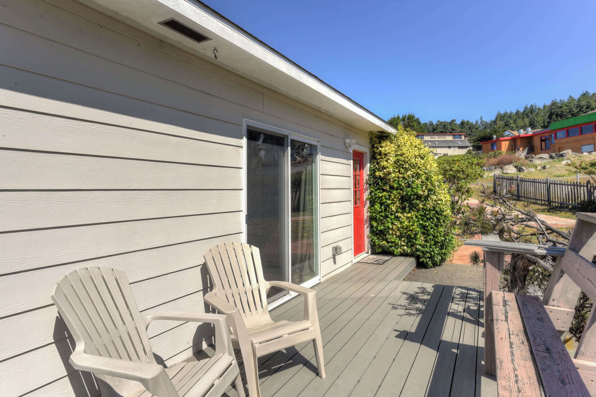 A wooden deck with two white plastic chairs, a red door, and a small garden area with a bush and a wooden fence.