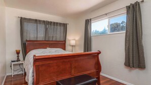 A bedroom with a wooden bed, a bedside table with a lamp, and a window with curtains. - Lucky Ewe - Gualala | 3BR | Ocean Views | Sleeps 6 (Gualala)