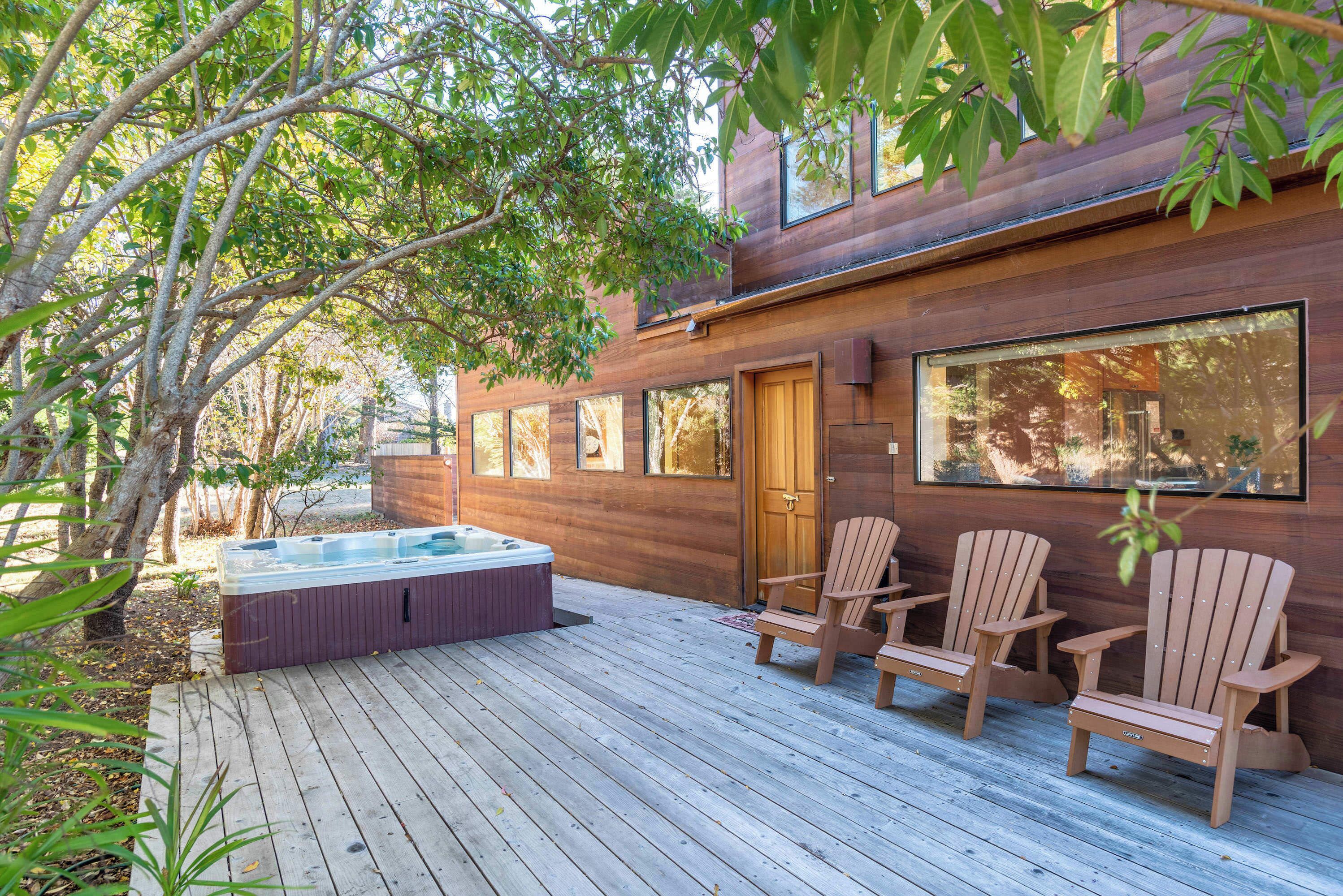 A wooden deck with a hot tub and Adirondack chairs, surrounded by trees.