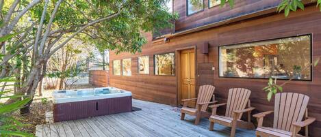 A wooden deck with a hot tub and Adirondack chairs, surrounded by trees.