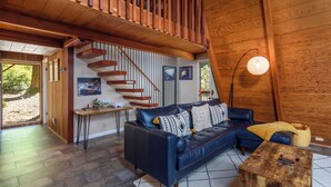 Een ruime woonkamer met een donkerblauwe leren bank, houten vloeren, een trap met houten leuningen en een groot raam met uitzicht op de omgeving.