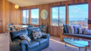 A modern living room with a leather sofa, a glass coffee table, and a dining area with a wooden table and chairs. There is a large window providing a view of the outdoors. - 3BR for 6 | Near Beach | Hot Tub | Ocean Views (Sea Ranch)