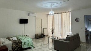 Uma sala de estar moderna com um sofá, uma TV na parede, uma área de jantar com mesa e cadeiras, e uma janela grande com cortinas.
