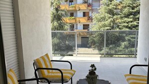 Uma varanda com quatro cadeiras listradas, uma pequena mesa com uma planta em vaso e vista para um edifício residencial com varandas e árvores.