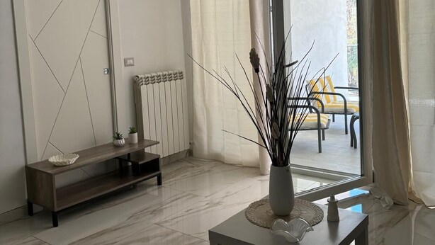 Uma sala de estar moderna com piso de mármore, um console minimalista, um radiador e vista para área externa com mesas e cadeiras através de uma porta de vidro.