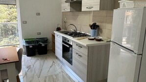 Cozinha moderna com fogão a gás, forno e frigorífico. Há uma mesa de jantar com cadeiras e uma janela com persianas.