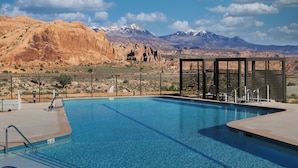 Una piscina con montañas al fondo, un área cercada y una zona de estar con sombra.