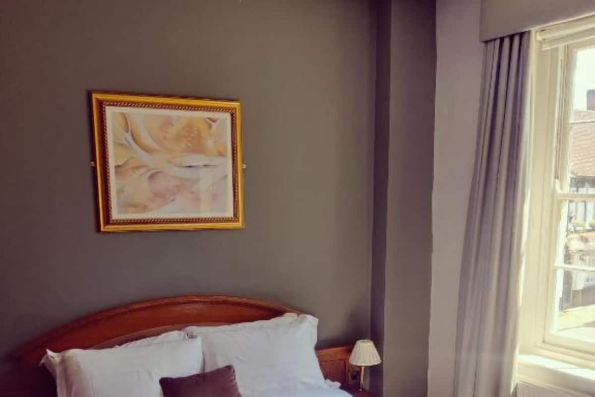 A neatly made bed with white linens, a wooden headboard, and a single brown pillow. A framed artwork hangs on the wall above the bed.