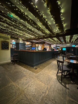 Een bar met een toonbank, krukken en tafels. Een bordje met 'CHECK IN' en een bordje 'TOILETS' zijn zichtbaar.