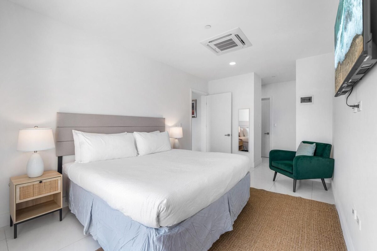 A modern hotel room with a large bed, bedside lamps, a green armchair, and a wall-mounted TV.