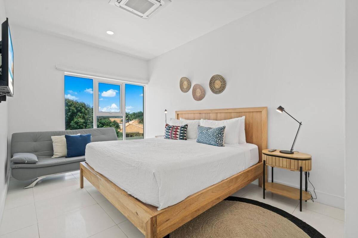 A bedroom with a large bed, a grey sofa, a desk with a lamp, and a TV on the wall.