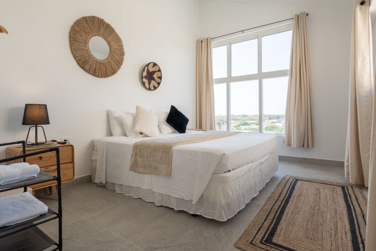 A bedroom with a bed, a bedside table with a lamp, a window with curtains, and a view of the outside.