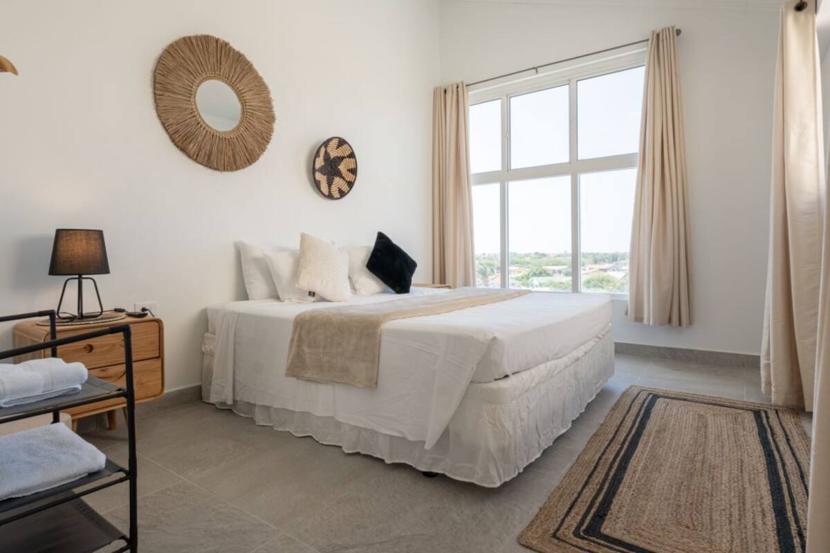 A bedroom with a bed, a bedside table with a lamp, a window with curtains, and a view of the outside.