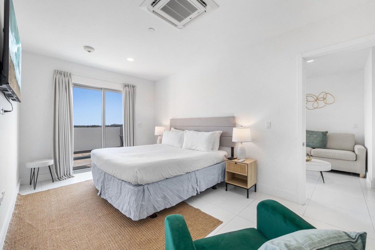 A modern hotel room with a large bed, bedside lamps, a small table, a green armchair, and a view of the outdoors.