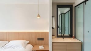 Een moderne hotelkamer met een houten hoofdbord, een nachtkastje met lamp, een grote spiegel, en een minimalistische badkamer met een douchecabine van glas.