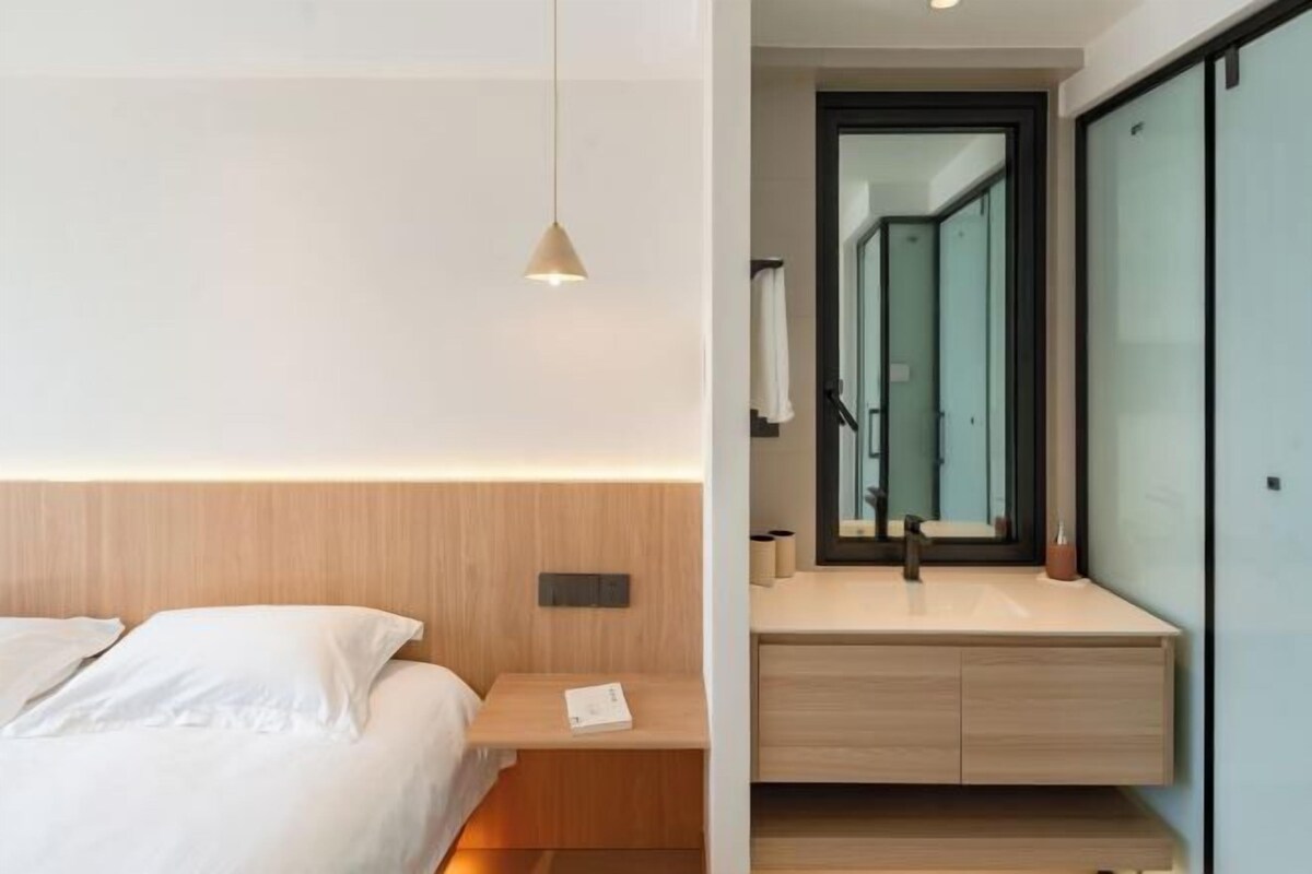 A modern hotel room with a wooden headboard, a bedside table, a nightstand, a built-in desk with a mirror, and a large window.