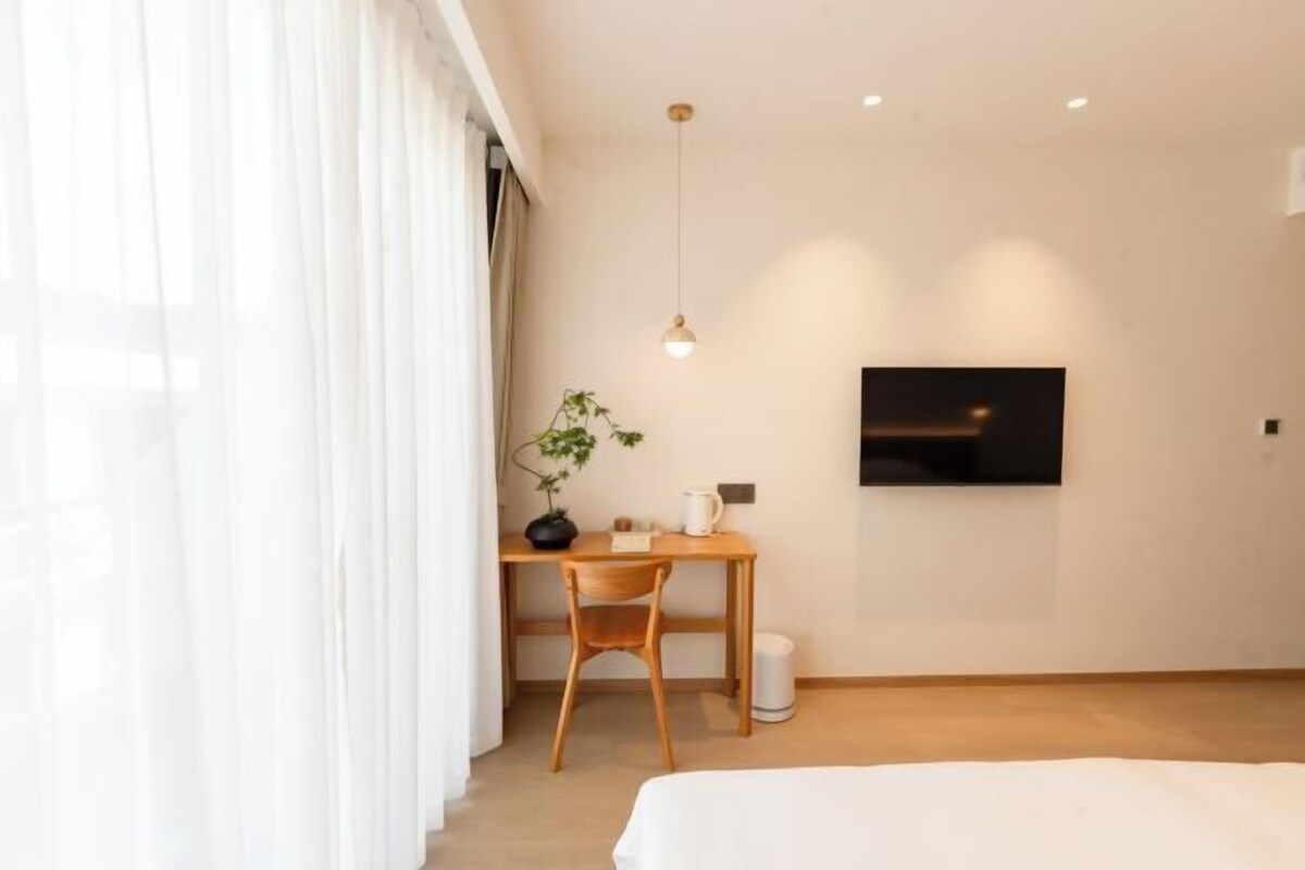 A modern hotel room with a wooden desk, a chair, a potted plant, a television mounted on the wall, and a bed with a white comforter.