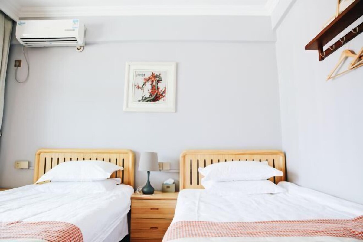 A hotel room with two single beds, a wall-mounted air conditioner, a framed artwork, and a wooden shelf with hangers.