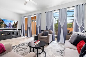A spacious living area with a sofa, armchair, and coffee table, a television, and a balcony with curtains.