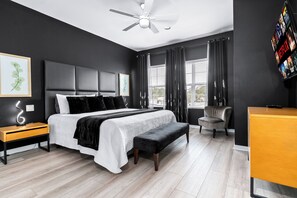 A modern bedroom with a bed, bedside tables, a desk, a chair, a ceiling fan, and a TV mounted on the wall.