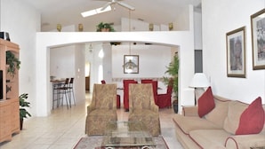 Een ruime woonkamer met een hoge plafond, een glazen salontafel, een bank en een eethoek met rode stoelen.