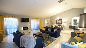 A modern living room with a fireplace, a TV mounted on the wall, and a dining area in the background. - Hummingbird Haven (Davenport)