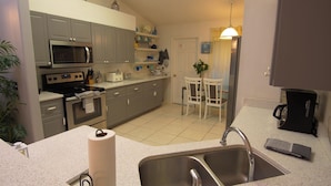 A kitchen with grey cabinets, a microwave, stove, and a coffee machine. There is a sink with a towel and a roll of paper towels. - Hummingbird Haven (Davenport)