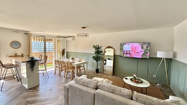 Een moderne woonkamer met een bank, eettafel en stoelen. Er is een televisie aan de muur en een potplant.