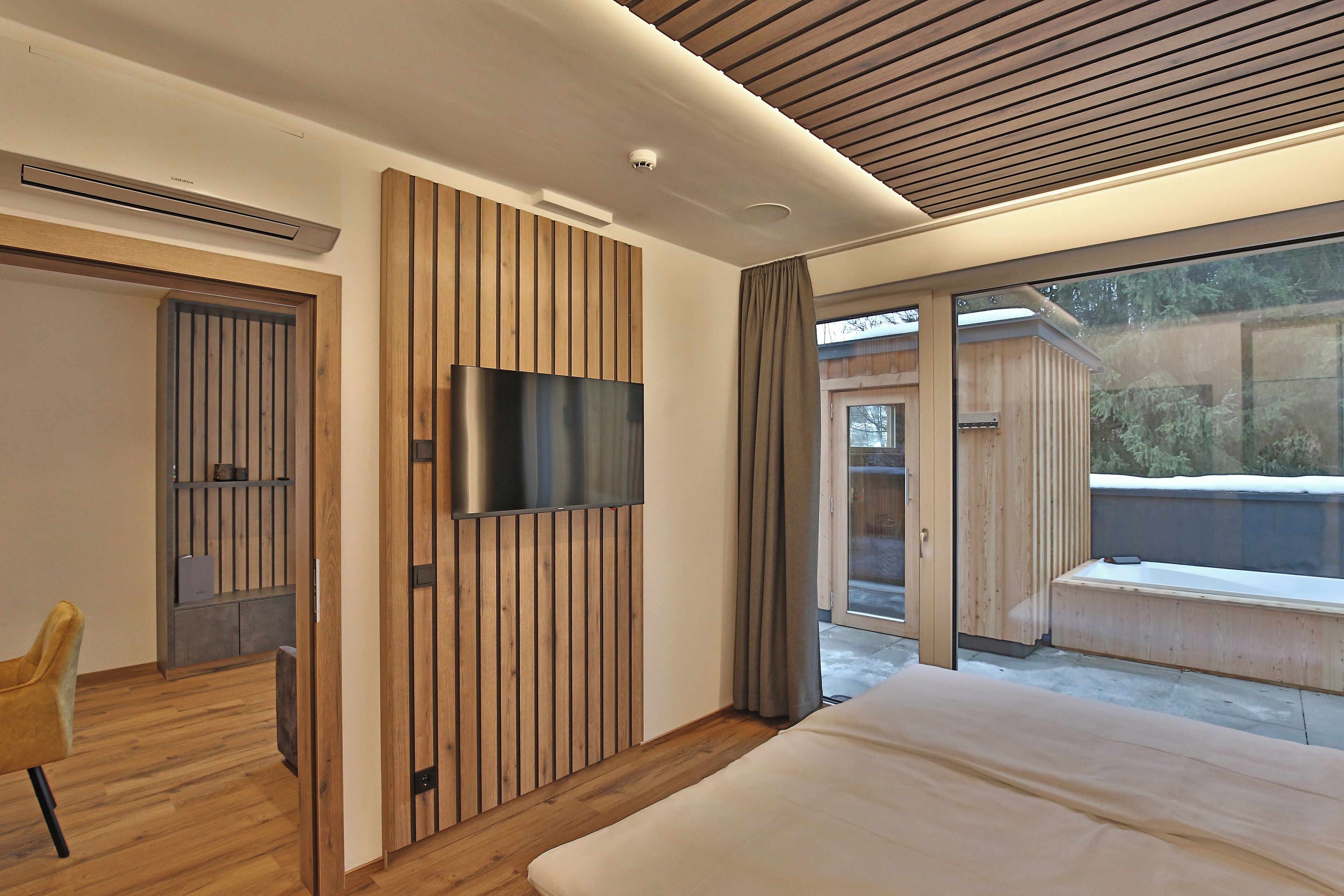 A modern bedroom with a large bed, a TV mounted on a wooden panel wall, and a sliding glass door leading to a balcony.