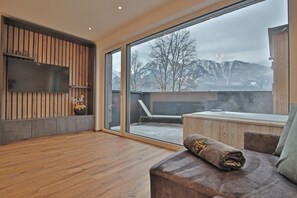 A modern room with a wooden floor, a large sliding glass door leading to a balcony, a flat-screen TV mounted on a wooden wall, and a cozy seating area with a cushioned bench and a rolled-up blanket. - Das Goldjuwel (Goldegg)