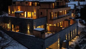 Un edificio de varios pisos de madera, con ventanas iluminadas y un techo cubierto de nieve, rodeado de árboles y otros edificios.