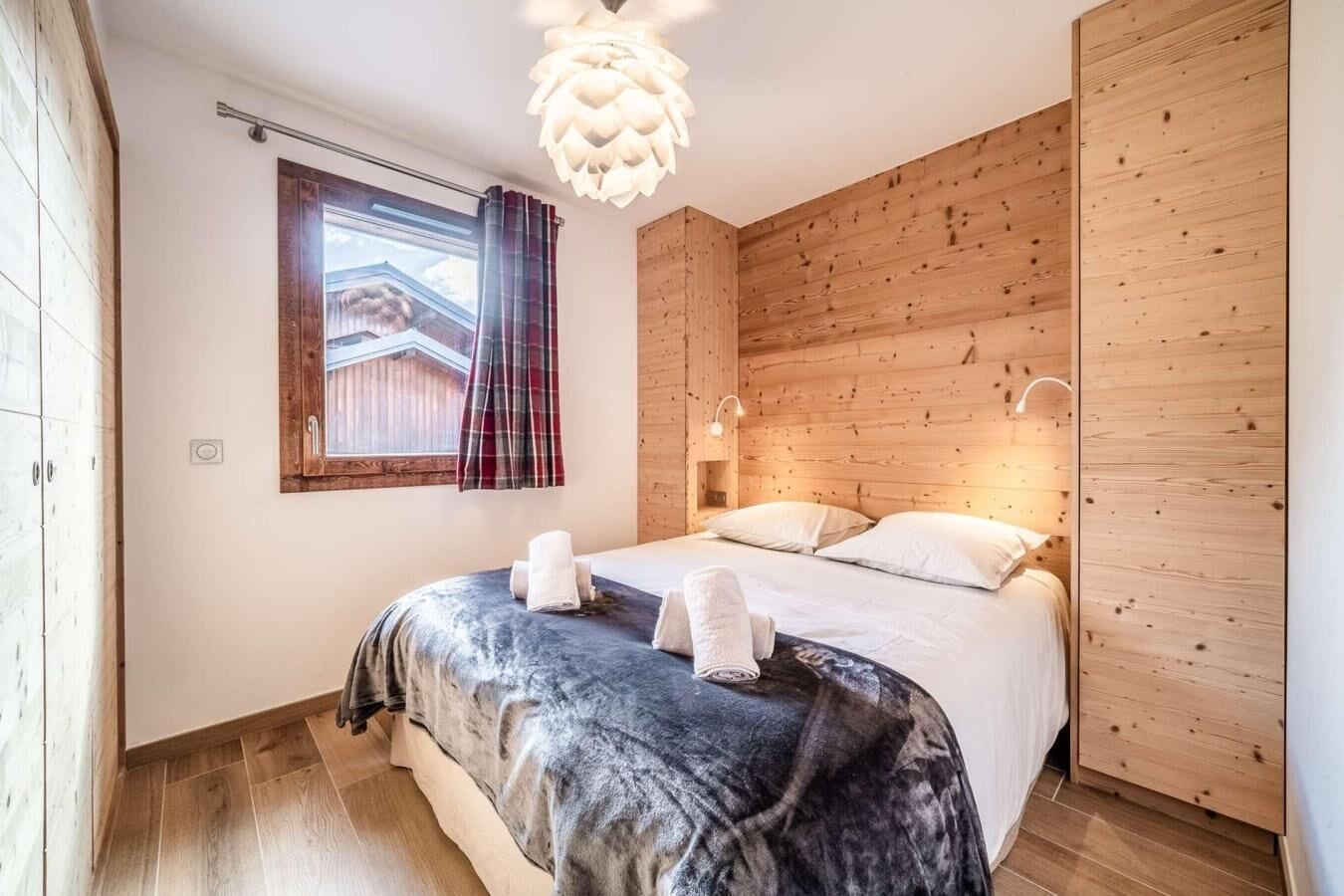 Un dormitorio con cabecera de madera, cama grande con colcha estampada oscura, dos almohadas y una ventana con cortinas a cuadros.