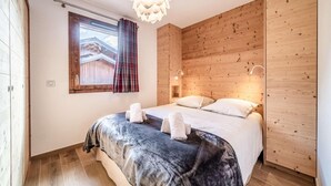 A bedroom with a wooden headboard, a large bed with a dark patterned bedspread, two pillows, and a window with plaid curtains.