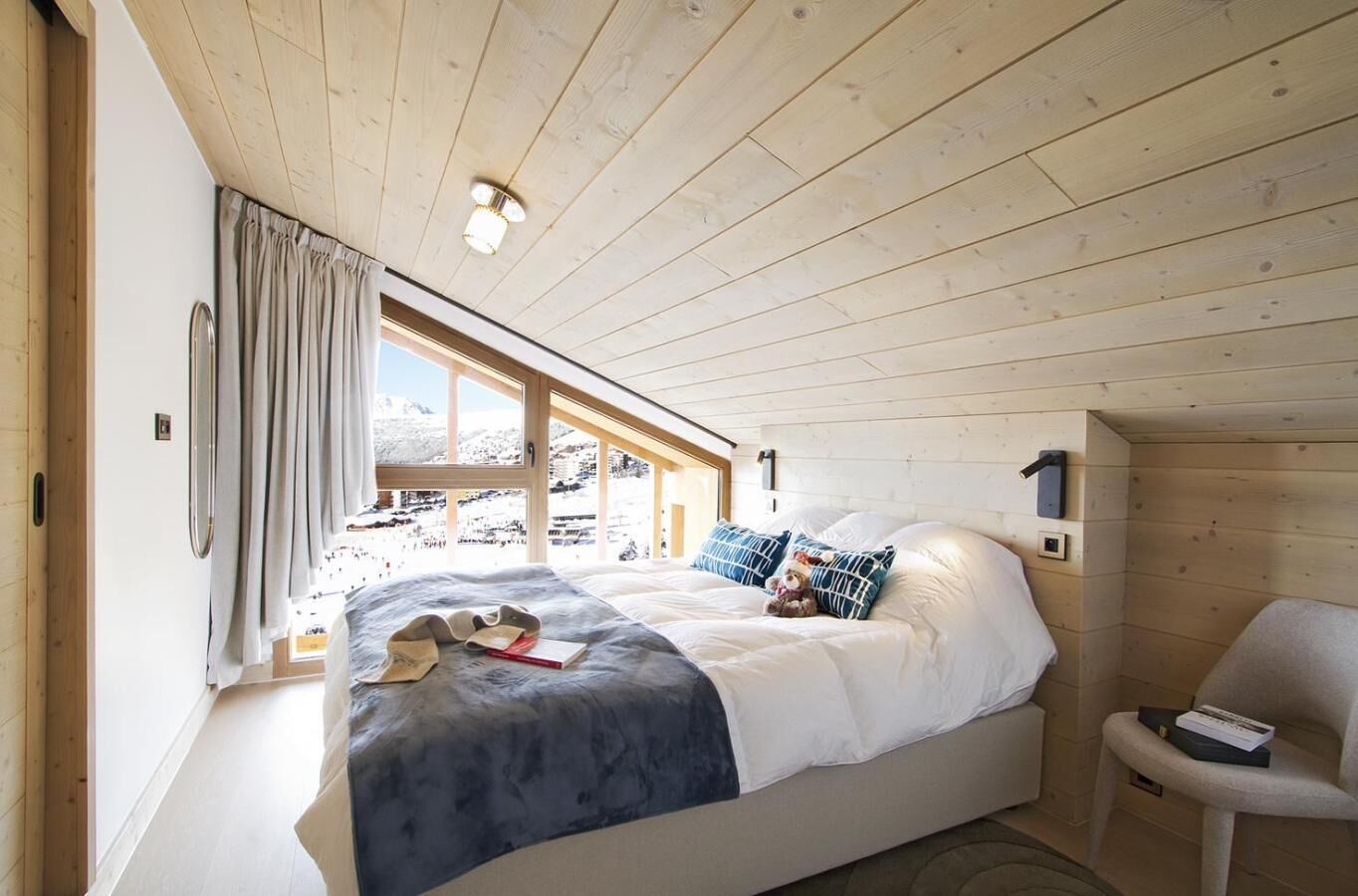 A bedroom with a bed, a chair, a window with curtains, and a wooden ceiling.