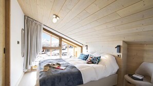 Ein Schlafzimmer mit einem Bett, einem Sessel, einem Fenster mit Vorhängen und einer Holzbalkendecke.