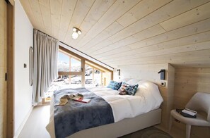 A bedroom with a bed, a chair, a window with curtains, and a wooden ceiling.