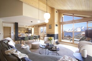 Sala de estar moderna com uma grande janela que oferece vista para as montanhas, um espaço de estar acolhedor e uma área de refeições com cozinha ao fundo.