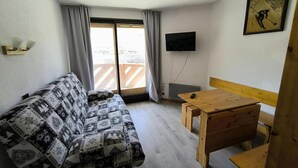 A small, furnished room with a sofa, a wooden table, and a TV mounted on the wall.