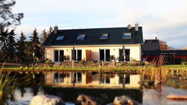 A modern house with a large front porch, a pond in the foreground, and a forested area in the background. - Naturferienhof Märkisch Luch - Trapphenne (Märkisch Luch)