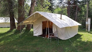 A tent with a single chair inside, set up in a grassy area with trees in the background. - Trapper Wall Tent, Trapper Wall Tent by the Stream (Story)