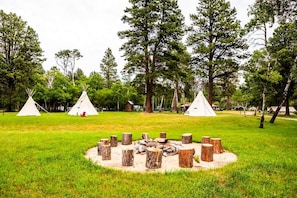 Un camping con tres tiendas de campaña blancas, un brasero central rodeado de troncos de madera y un espacio cubierto de césped con árboles al fondo.