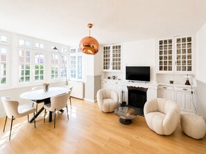 A modern living room with a fireplace, a dining area with a round table and chairs, and large windows with white frames.