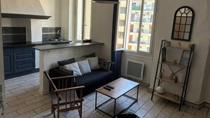 A compact living space with a kitchenette, a sofa, a coffee table, and a window with a view of a balcony. - Bright Apartment Near the Beach and Old Port (Marseille)