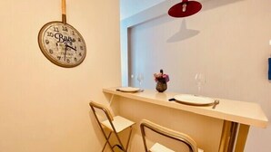 Ein kleiner Essbereich mit Wanduhr, zwei Klappstühlen, einem Tisch mit Blumenvase und Weingläsern.