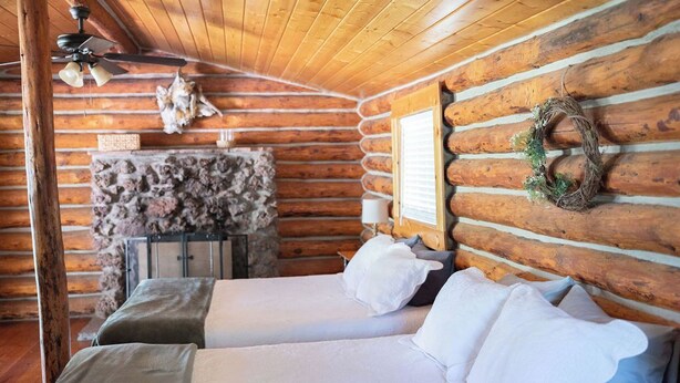 A log cabin bedroom with two beds, a stone fireplace, wooden walls, and a ceiling fan.