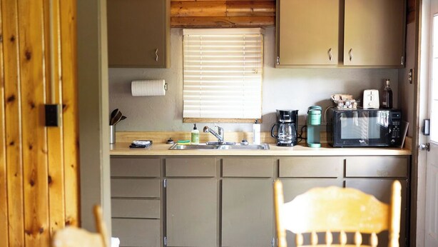 A compact kitchen with wooden cabinets, a sink, a window with blinds, and a microwave on the counter. - Creekside Chalet, Private Creekside Chalet (Story)