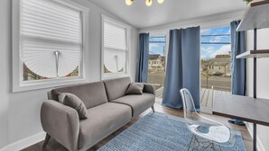 Un salón moderno con un sofá gris, una silla transparente, una mesa de madera y vistas a la calle a través de ventanas grandes con persianas blancas y cortinas azules.