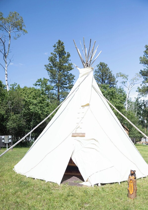A white teepee with a bear symbol on the entrance, set in a grassy area with trees in the background. - Bear Tipi, Rustic "Bear" Tipi by Bighorn Mountains (Story)